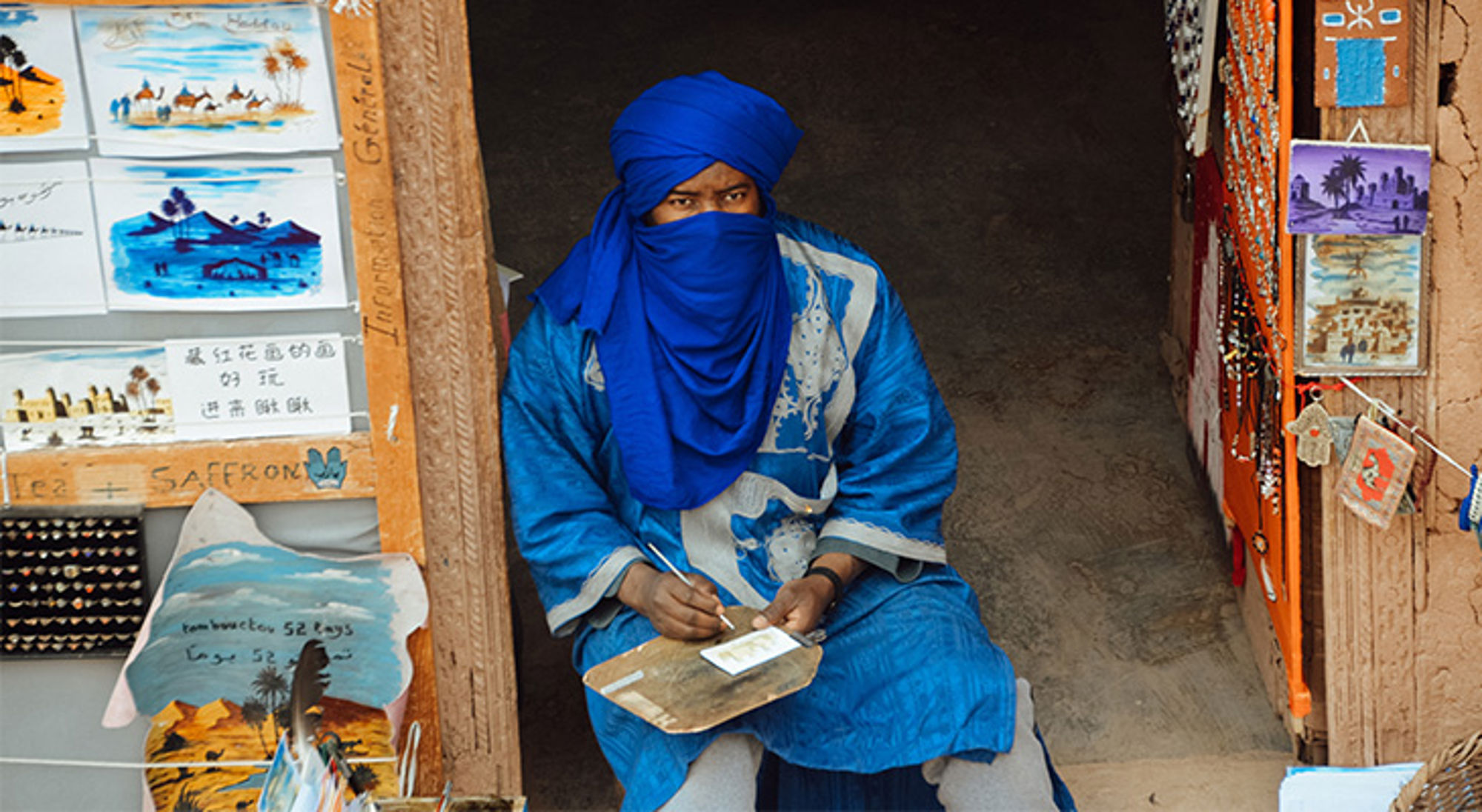 Kunstenaar in Aït-Ben-Haddou Man in blauwe tulband maakt een schilderij bij een souvenirwinkel in de medina.