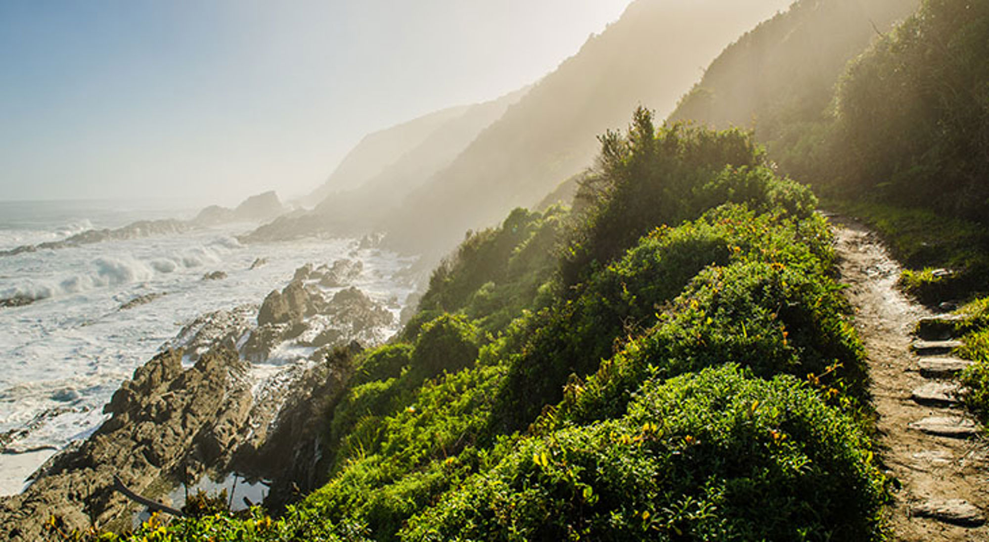 Garden Route Kustpad langs rotsachtige kust en groene heuvels in Zuid-Afrika, met uitzicht op de oceaan.