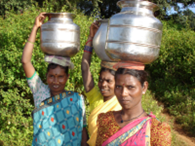 Vrouwen in Odisha dragen metalen waterkruiken op het hoofd langs een groen pad.