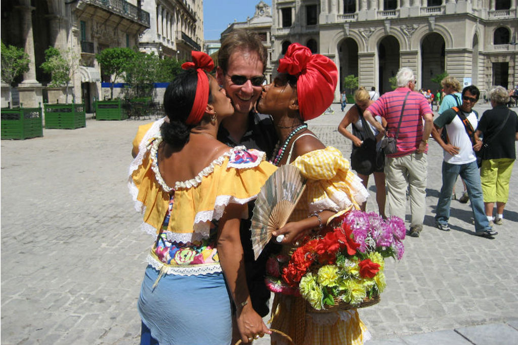 Twee vrouwen in kleurrijke kleding geven een toerist een kus op een plein in Havana, Cuba.