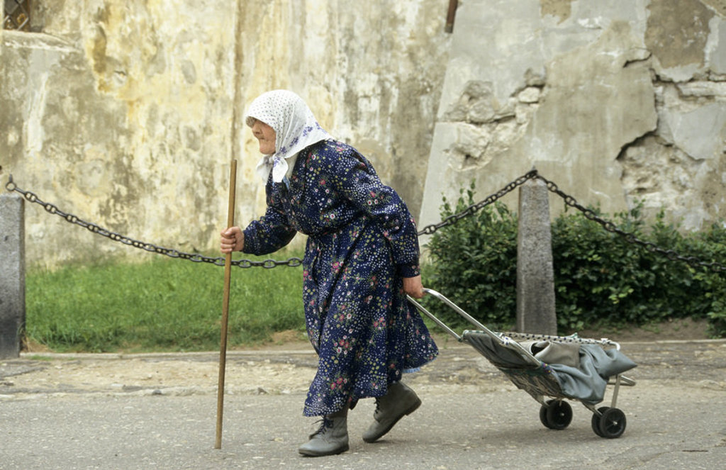 Oudere vrouw met hoofddoek en wandelstok trekt een karretje over straat in Rusland.