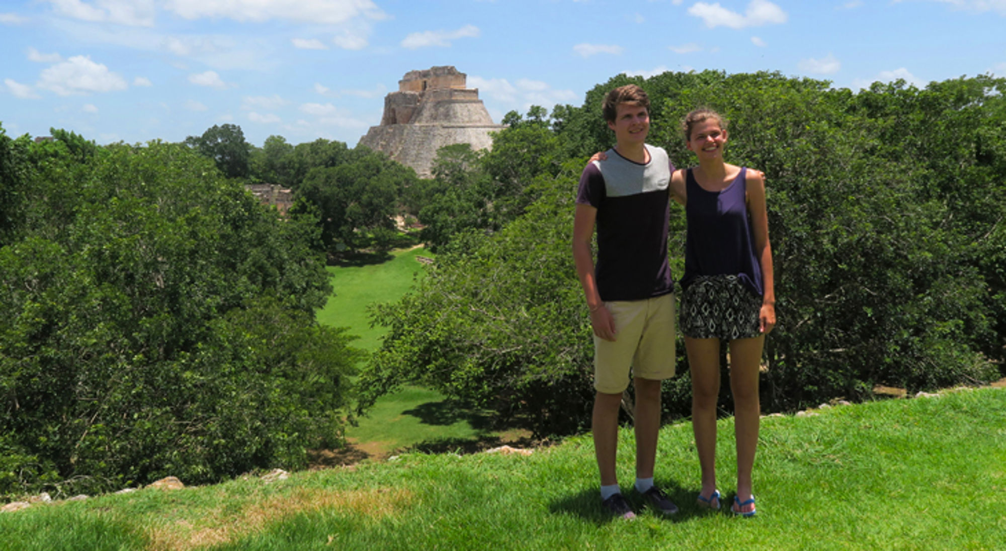 Twee reizigers poseren bij de Maya-piramide van Uxmal, omringd door groen en blauwe lucht.