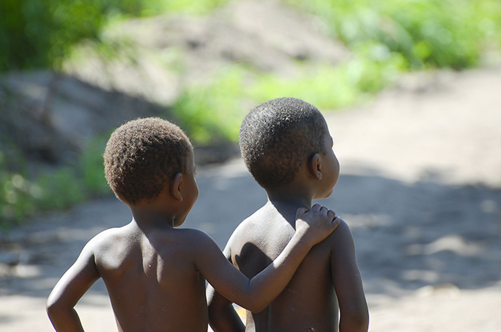 Twee kinderen lopen samen over een zandpad, één met een arm om de ander heen.