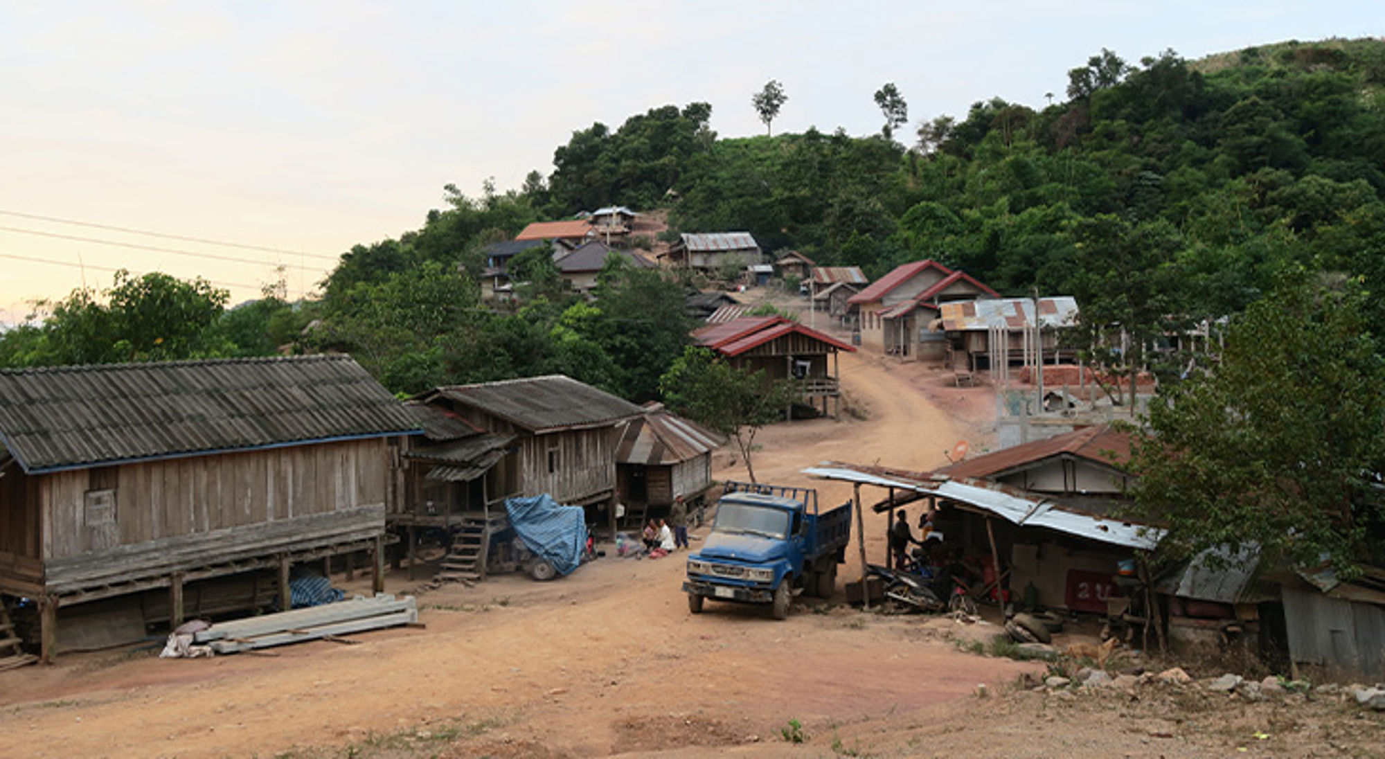 Bergdorpjes Noord-Laos Dorp met houten huizen langs een onverharde weg, met een blauwe vrachtwagen en groene heuvels op de achtergrond.