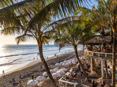 Zonsondergang op het strand van Bali met palmbomen, strandbar en surfers langs de kust.