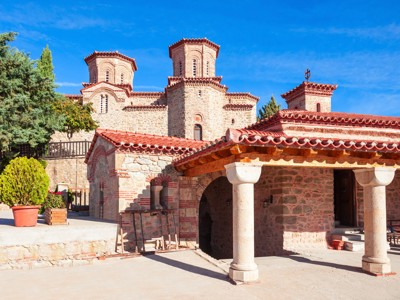 Binnenplaats van een klooster in Meteora, Noord-Griekenland, met stenen kerk en rode dakpannen onder blauwe lucht.