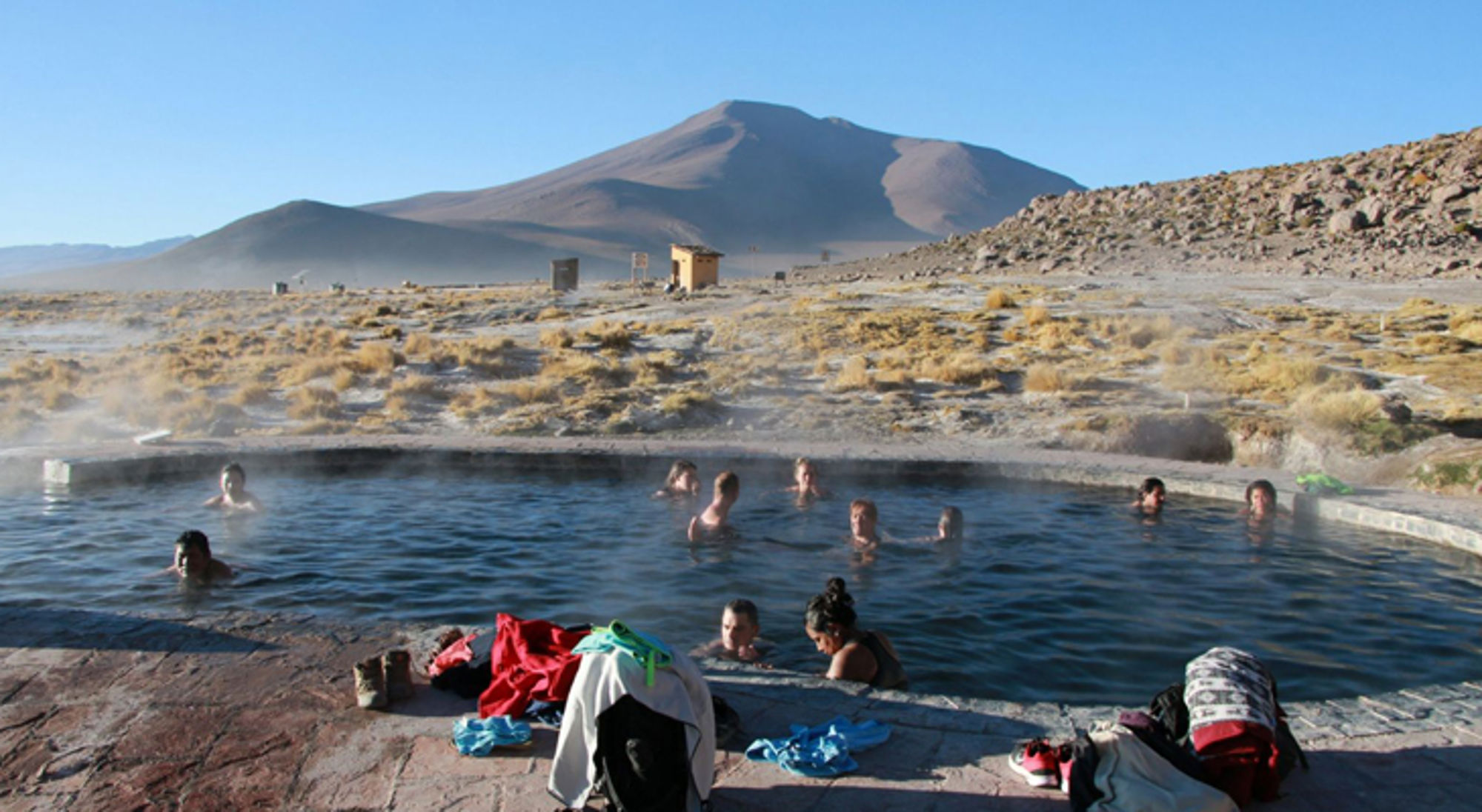 thermale baden in bolivia Badende reizigers in een warmwaterbron met uitzicht op een vulkaan in een droog berglandschap.