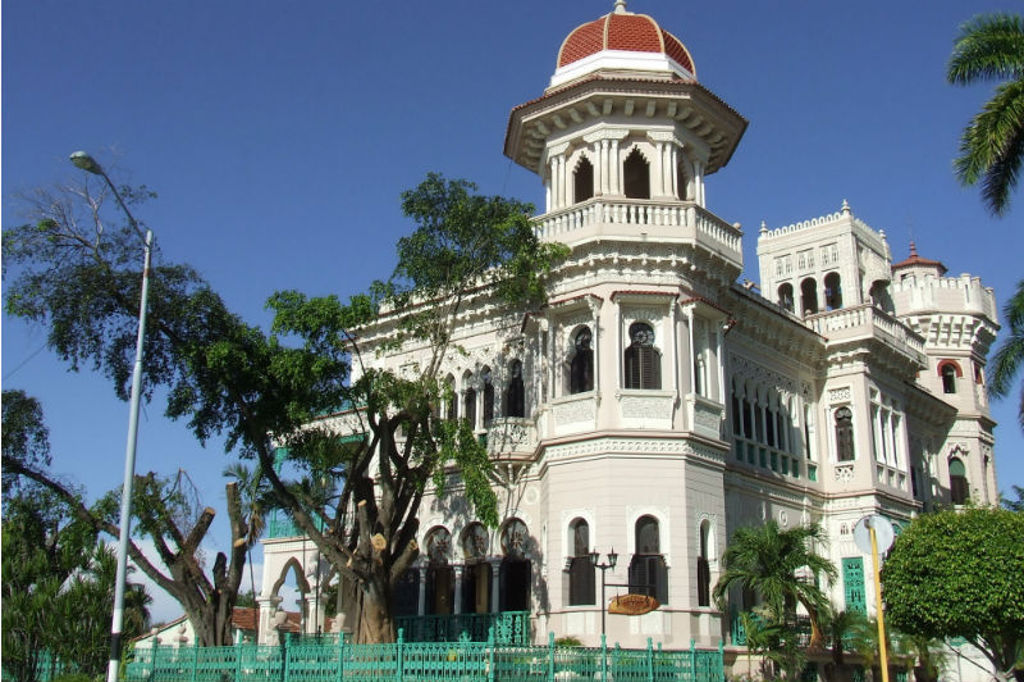 Wit historisch gebouw met toren en rode koepel in Cuba, omringd door palmen en groen.
